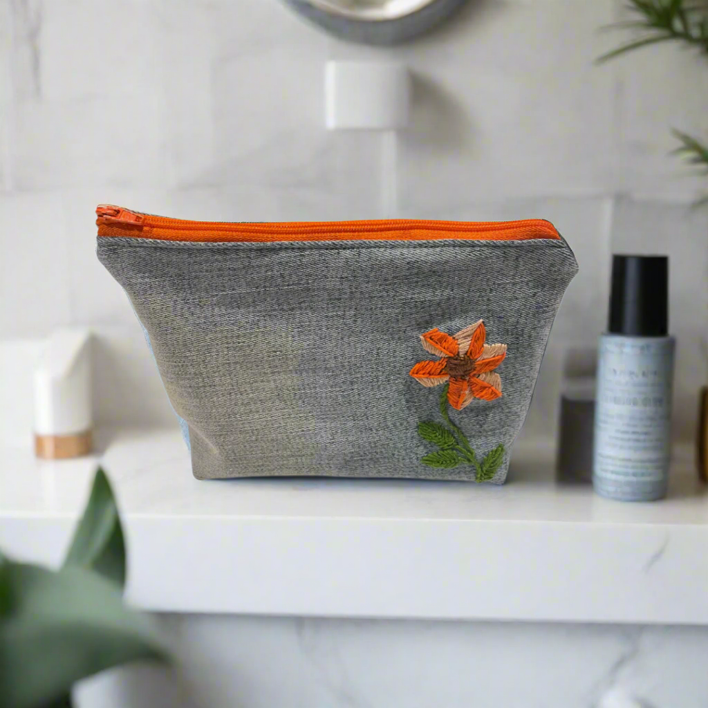 Light blue denim pouch with orange zipper and embroidered flower design on a white background