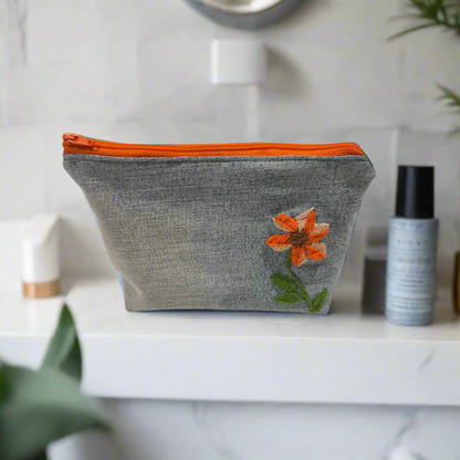 Light blue denim pouch with orange zipper and embroidered flower design on a white background