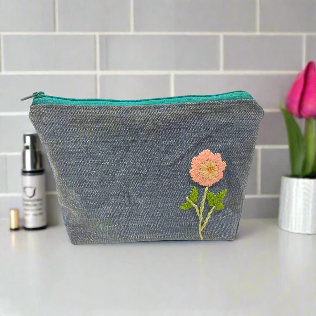 Blue denim pouch with floral embroidery on a table with makeup items and pink roses.